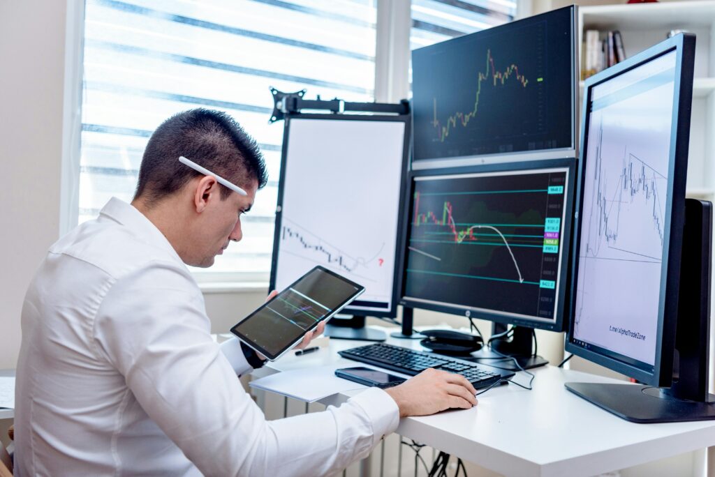 Man checking stats on a tablet while sat at a desk with 4 screens displaying graphs.